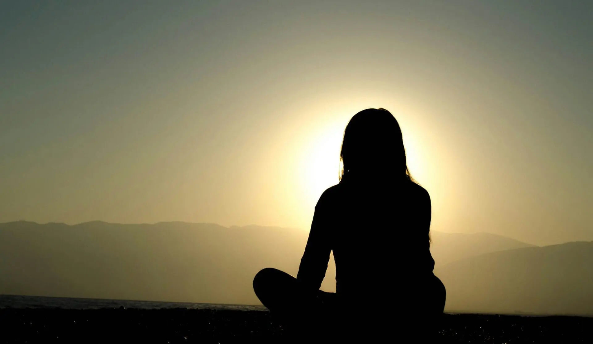 Silhouette of a person sitting cross-legged, facing a bright setting or rising sun, with mountains in the background.