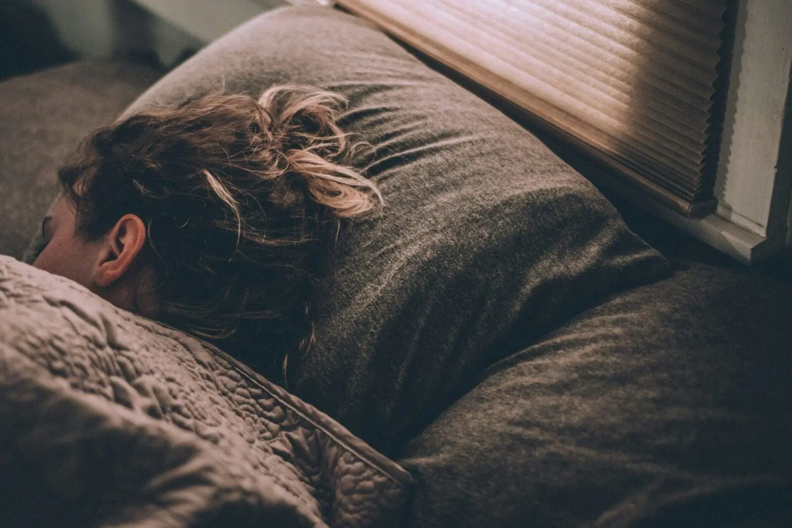 Person sleeping on a bed with a dark blanket and pillow, near a window with closed blinds.