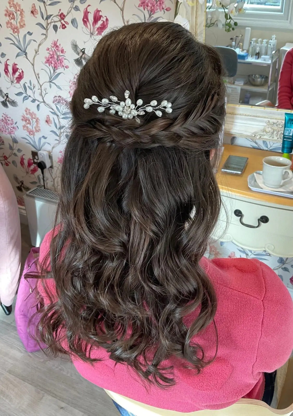Woman with long, wavy brown hair styled in a half-up braid, decorated with a pearl hairpiece, sits facing a vanity mirror. She is wearing a pink top.