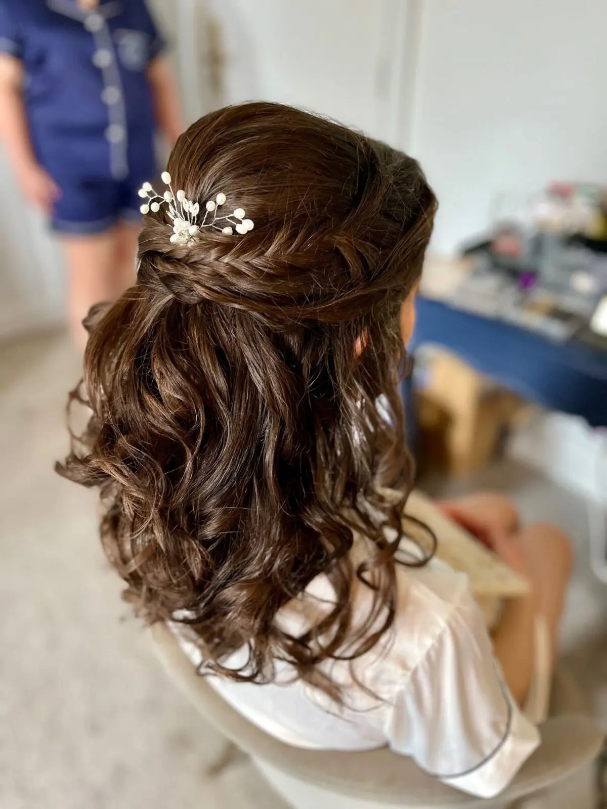 Woman with long, wavy brown hair styled in a half-up braid, adorned with a small hair accessory with white beads, sitting indoors.