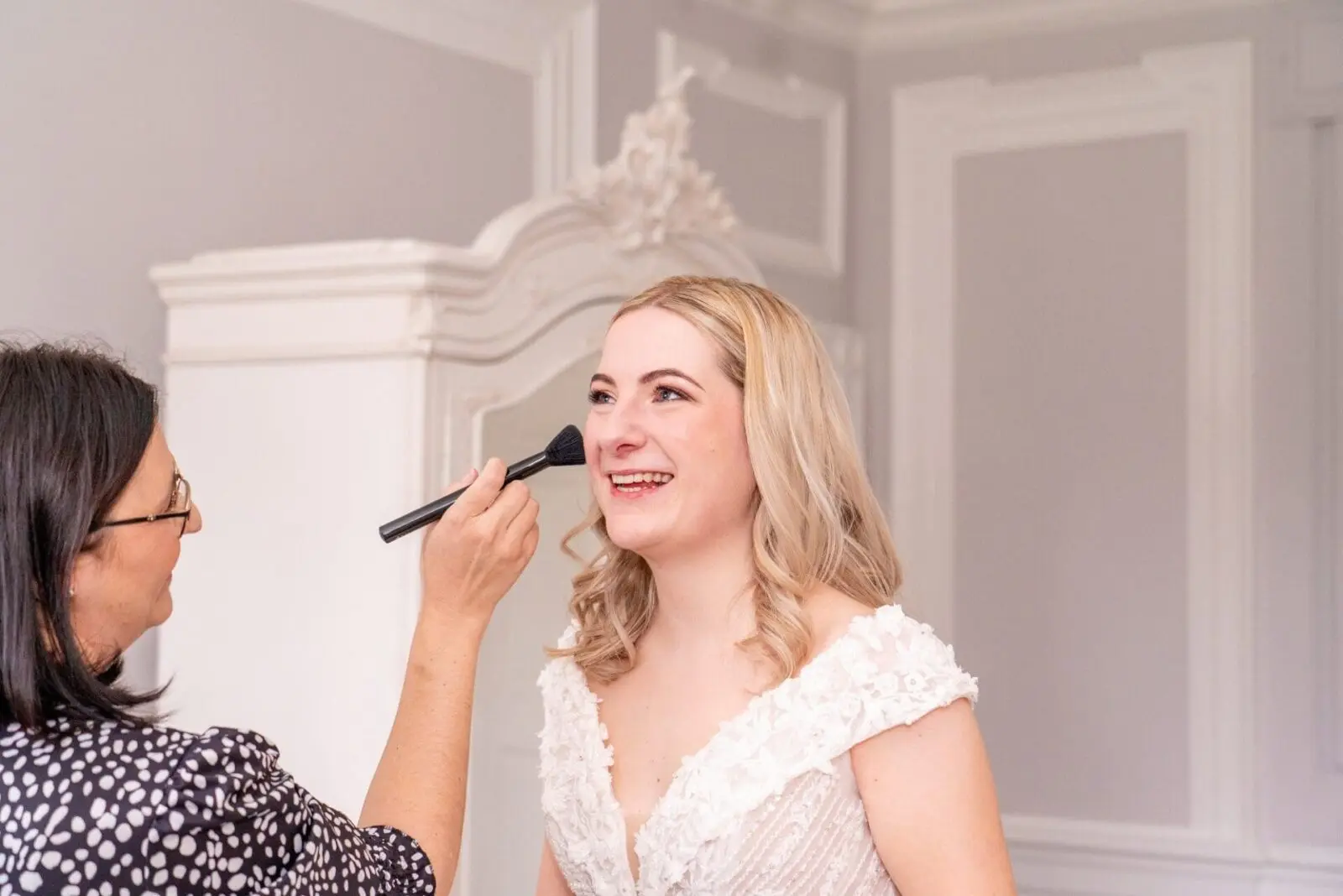 A woman in a white dress smiles as another person applies makeup to her face with a brush in a bright room.