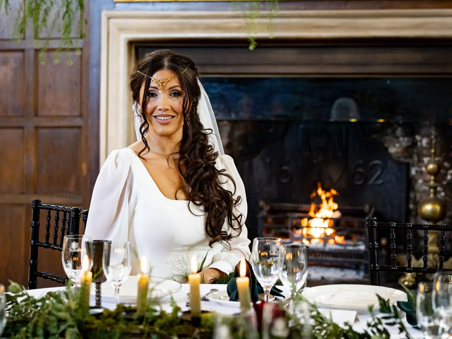 Bride in a white dress with long sleeves and headpiece sits at a table with lit candles and greenery, in front of a fireplace.
