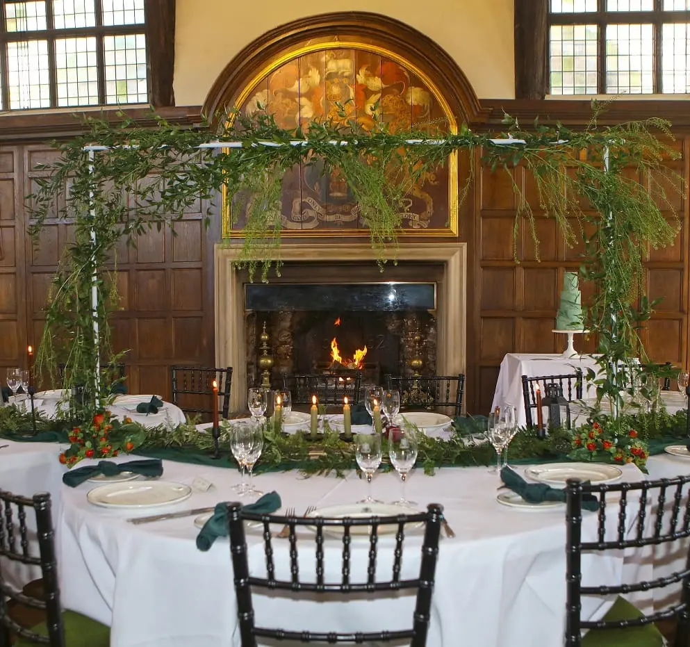 Elegant dining setup with round tables and black chairs in a wood-paneled room. A fireplace is lit, and greenery with candles decorates the tables. A green tiered cake is in the background.