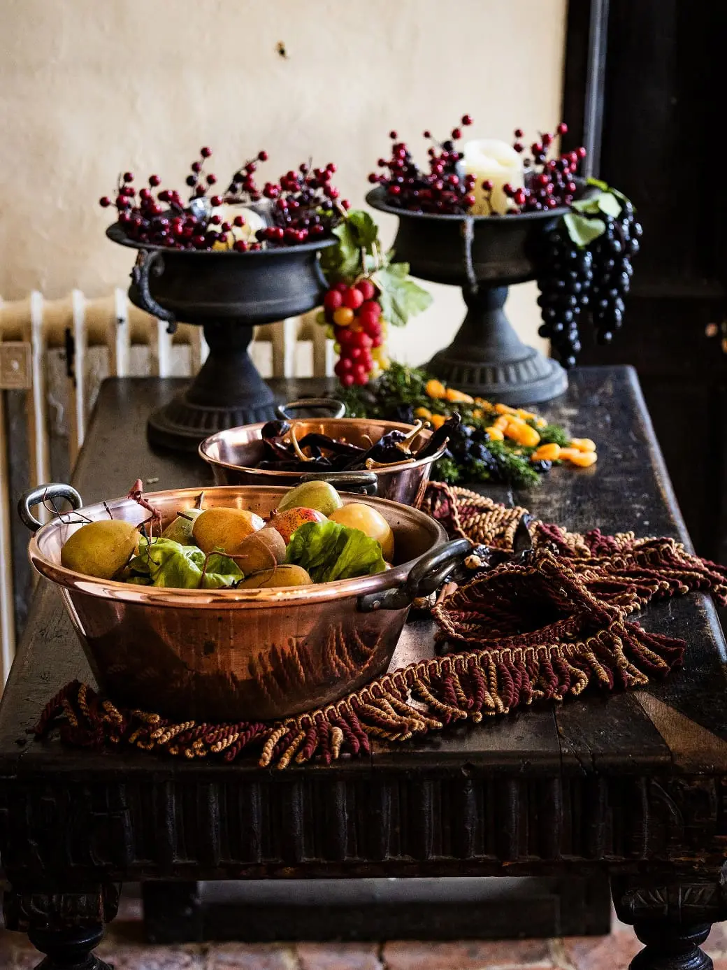 Copper bowls with apples and pears, and dark pedestals holding berries, grapes, and fruits on a wooden table, decorated with a textured cloth.