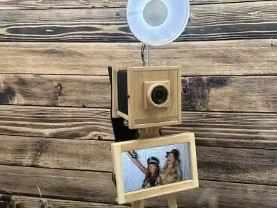 A vintage-style photo booth with a wooden exterior, camera lens, flash, and a screen displaying two people taking a selfie.