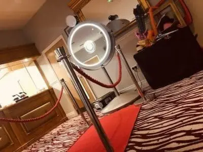 A photo booth ring light stands on a red carpet with stanchions, in a room with patterned carpeting and a mirror in the background.