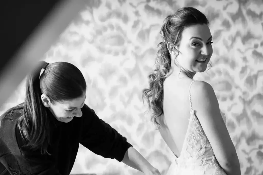 A woman helps a bride adjust her dress. The bride, wearing a lace gown, looks over her shoulder and smiles. The image is in black and white.