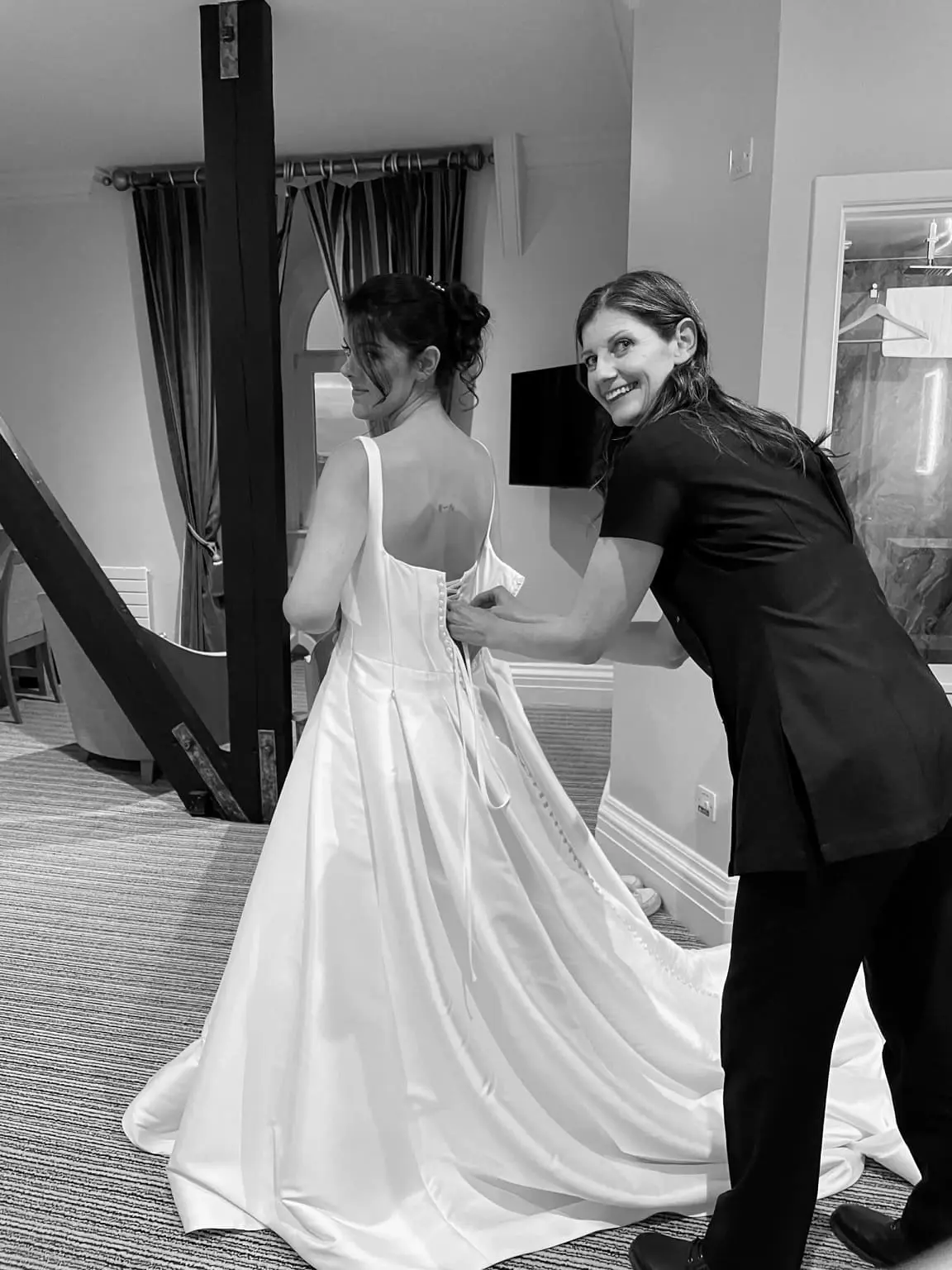 Two women are in a room; one is wearing a wedding dress while the other helps fasten the back of the dress. Both are smiling.
