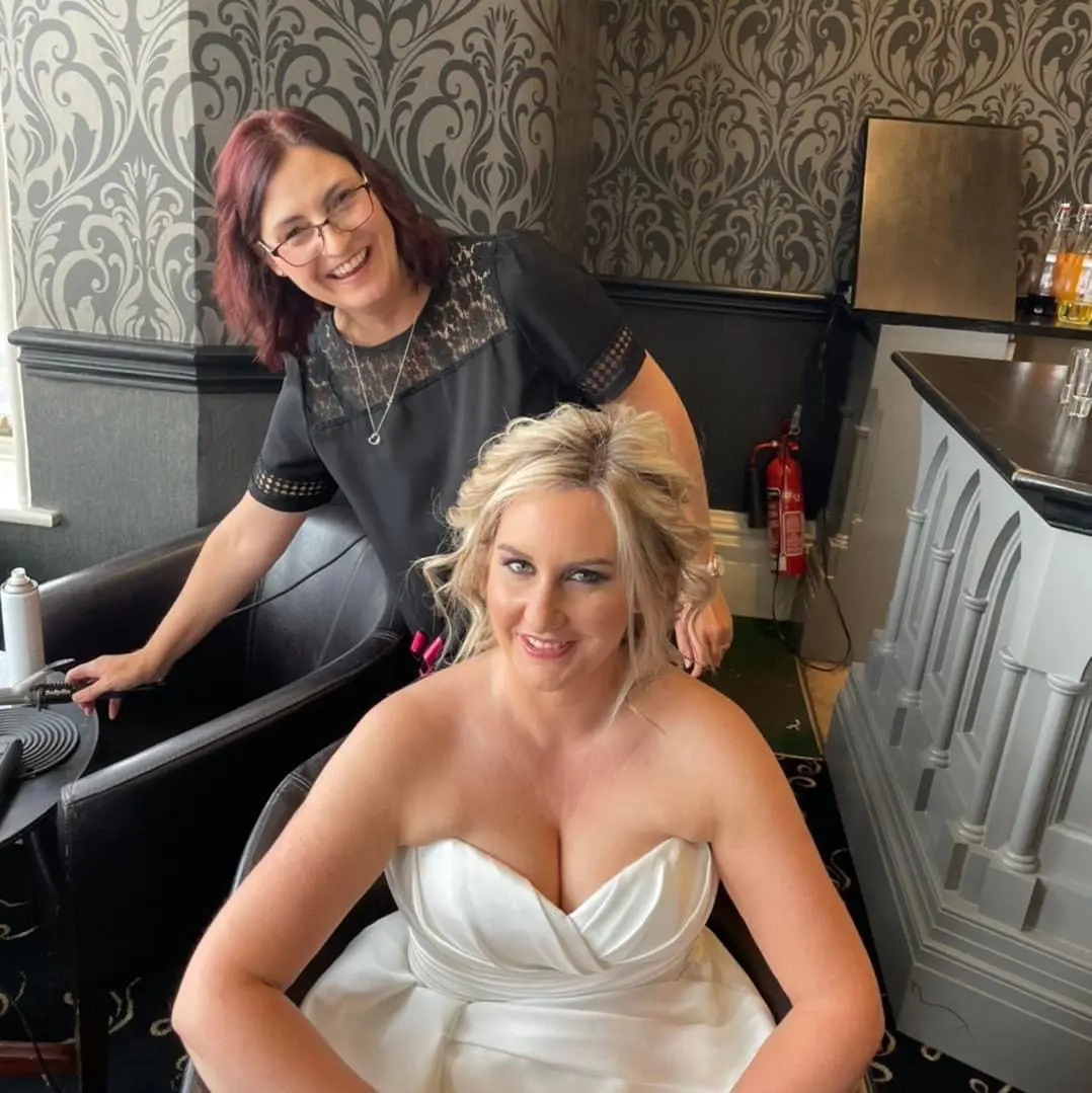 A woman in a white dress sits and smiles while another woman stands behind her, both indoors with patterned wallpaper and salon items visible.