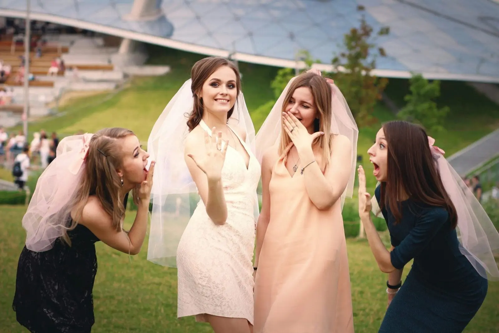 Four women, one in a white dress showing her ring, pose outdoors with veils; the others react excitedly, suggesting a bridal celebration.
