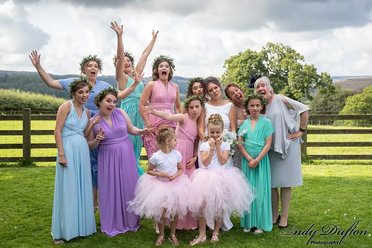 A group of women and young girls in colorful dresses and floral crowns pose playfully outdoors on grass with a wooden fence and trees in the background.