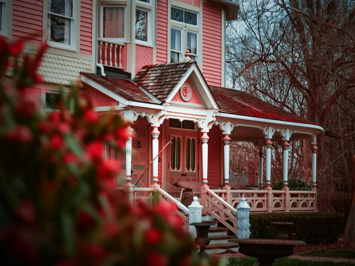 A large pink Victorian-style house with a covered porch and intricate white trim, partially obscured by red flowers in the foreground.