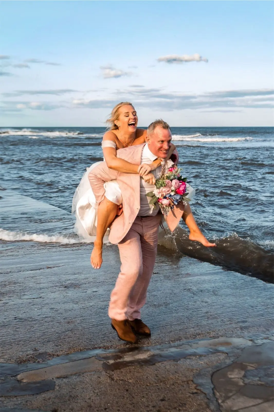 A man in a pink suit carries a woman in a wedding dress on his back along the shoreline, both smiling, with the sea and sky in the background.