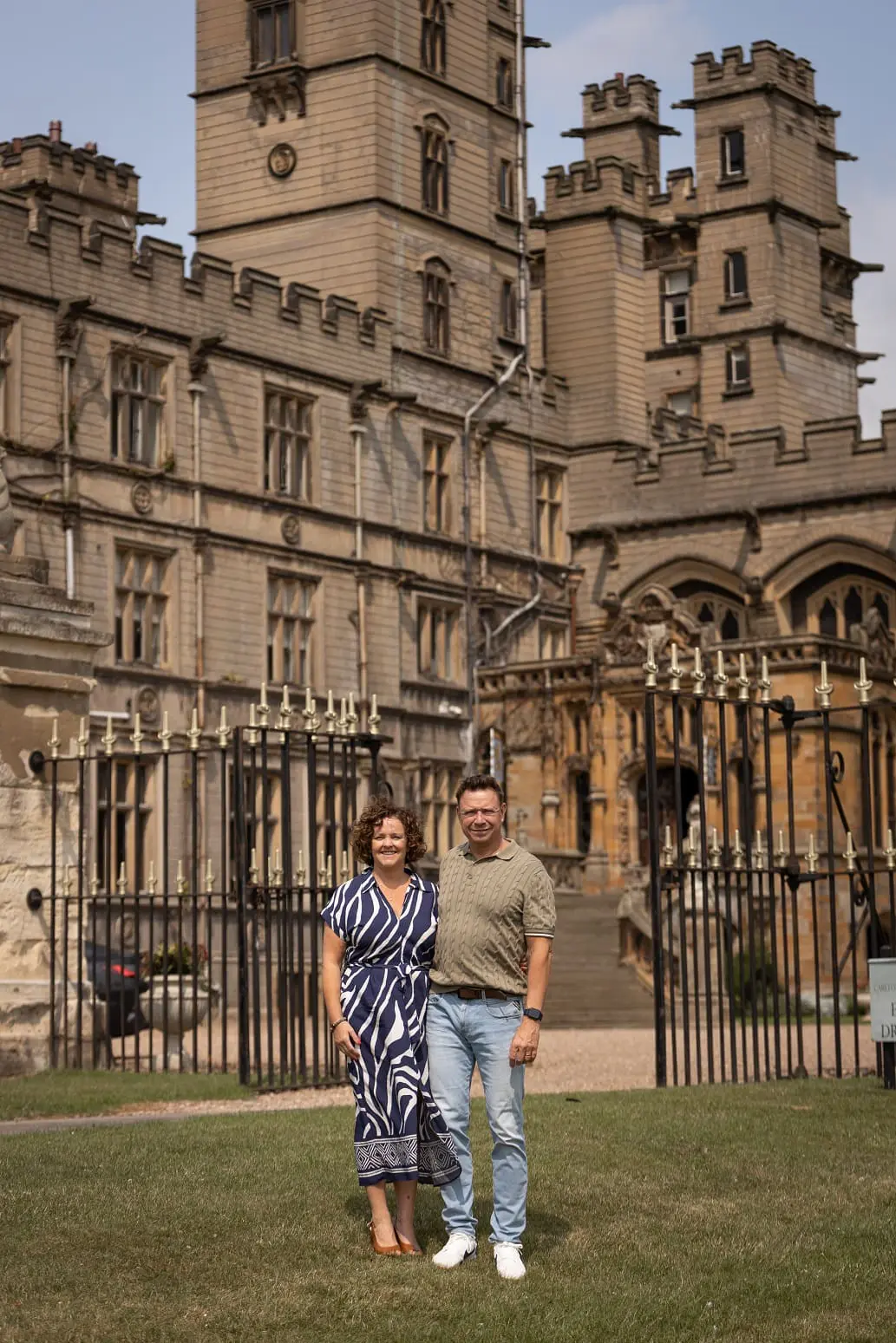 A man and a woman stand on grass in front of a large, historic stone building with ornate architectural details and iron gates.