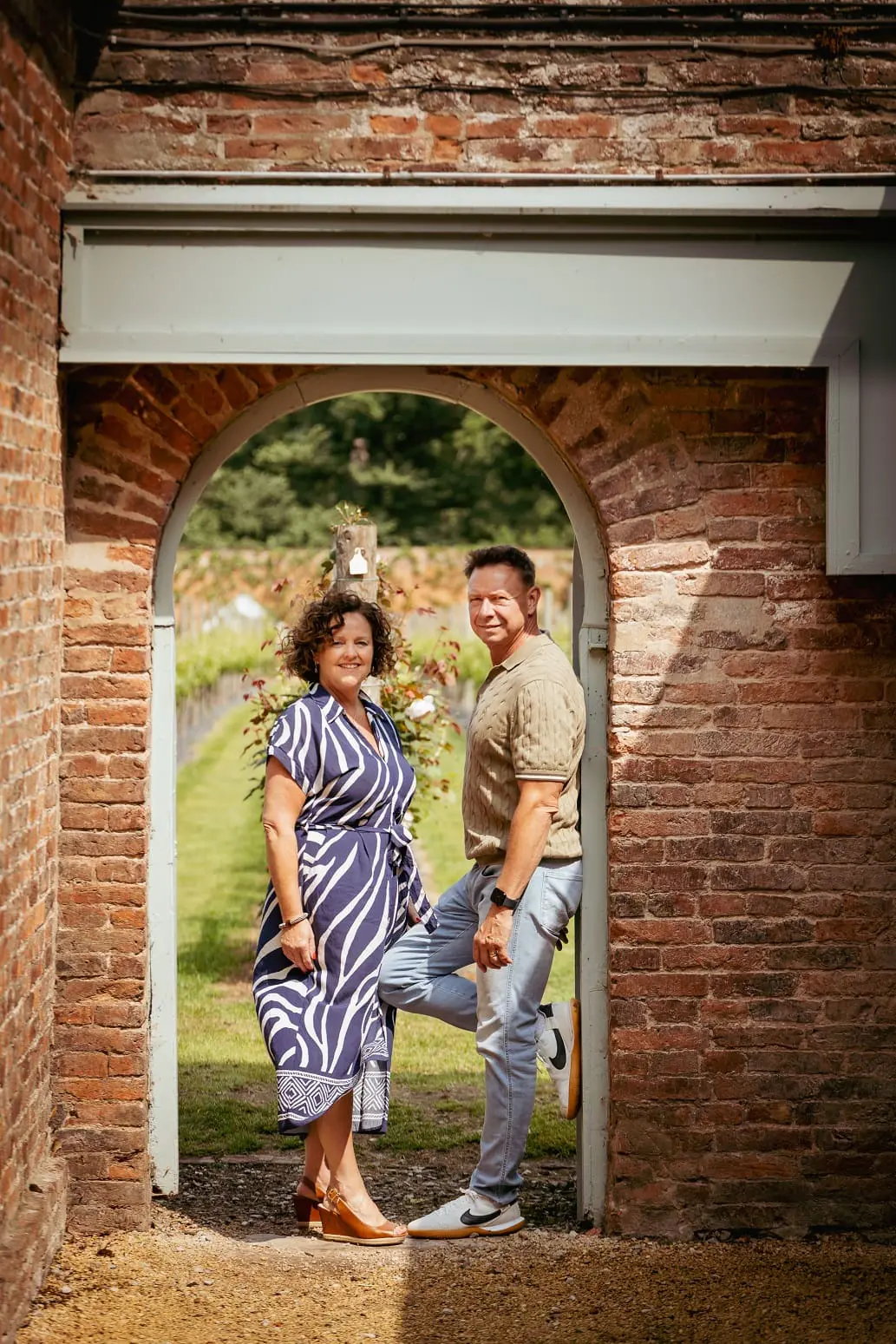 A woman and a man stand together under a brick archway in a garden, facing the camera. Greenery and flowers are visible in the background.