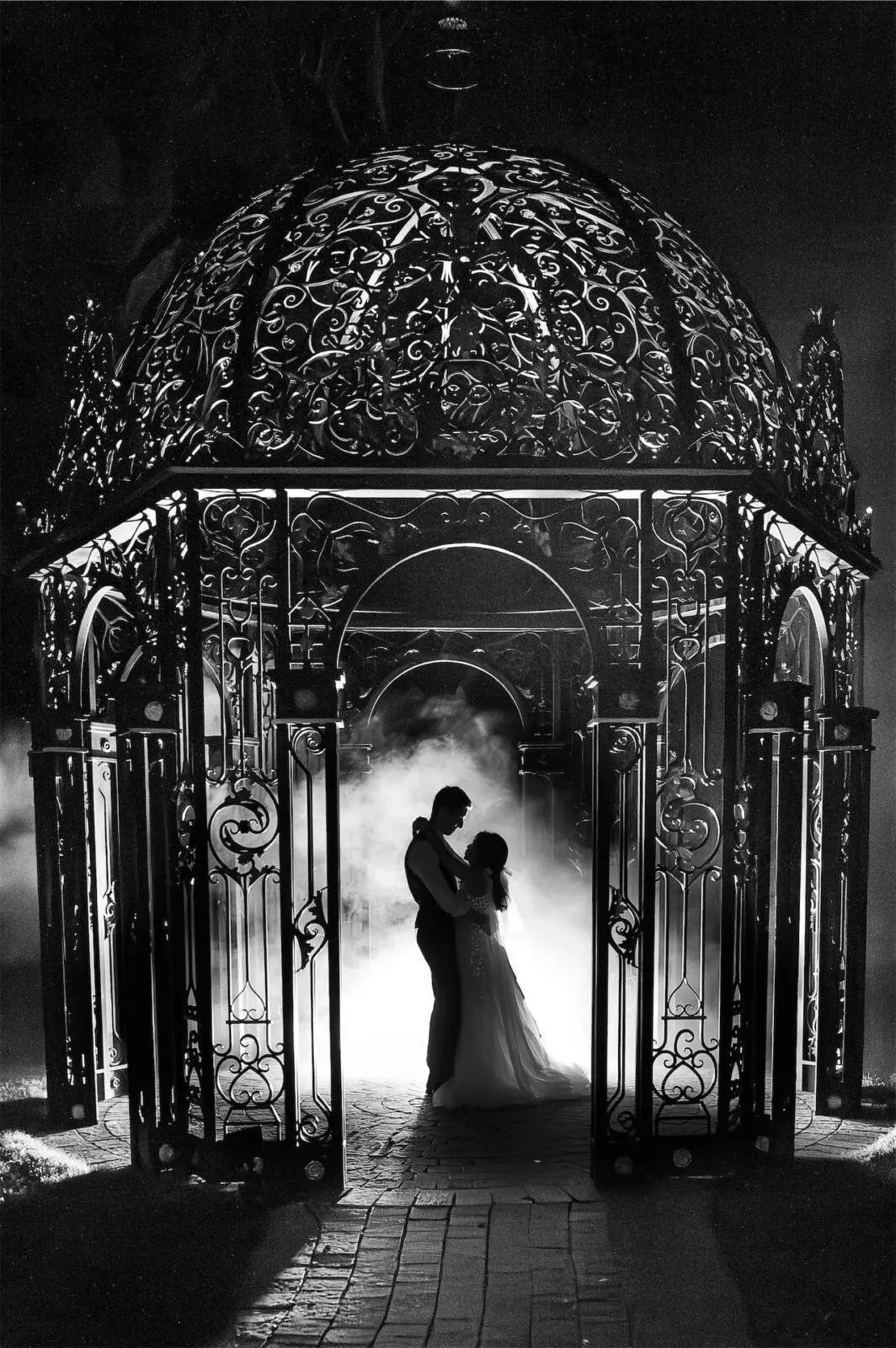 A couple stands embracing under an ornate, domed gazebo at night, with mist and dramatic lighting creating a romantic atmosphere.
