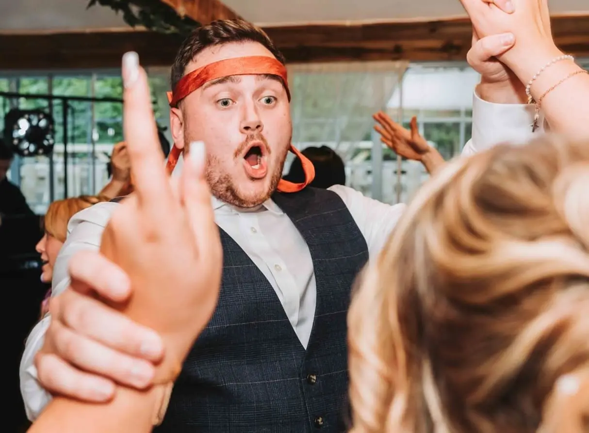 A man with a red ribbon around his head and an open-mouthed expression raises his hands while dancing at a lively indoor event.