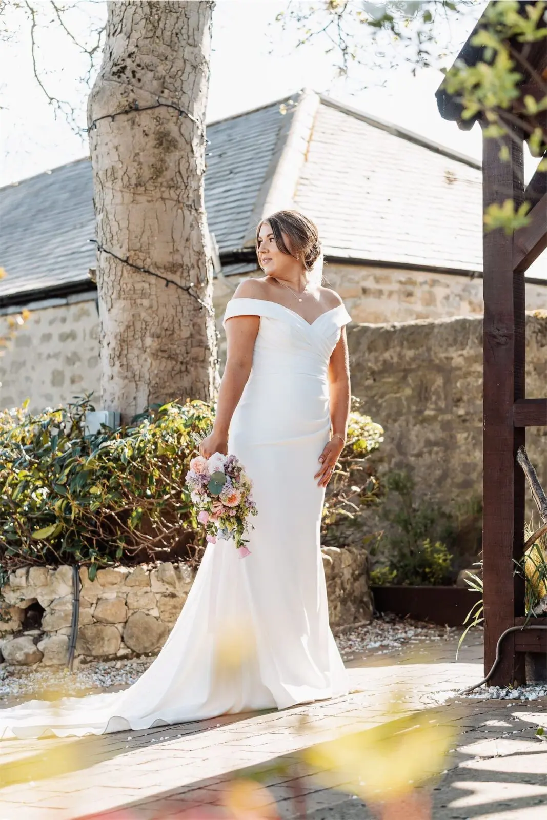 A woman in an off-the-shoulder white wedding dress stands outdoors, holding a bouquet of flowers, with stone buildings and greenery in the background.