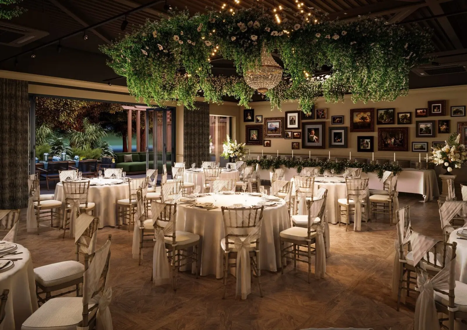 Elegant banquet hall with round tables and white chairs, greenery hanging from the ceiling, chandelier, framed art on the walls, and large windows opening to a garden.