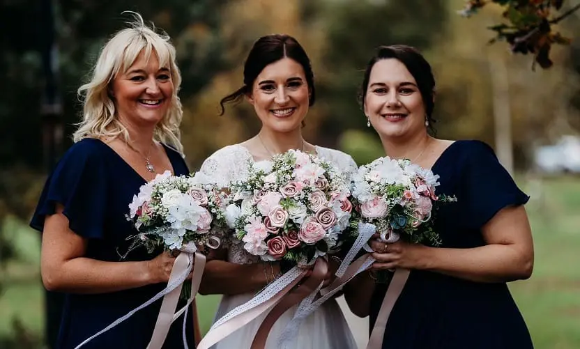 Three women in navy dresses holding pink and white flower bouquets, standing outdoors and smiling at the camera.