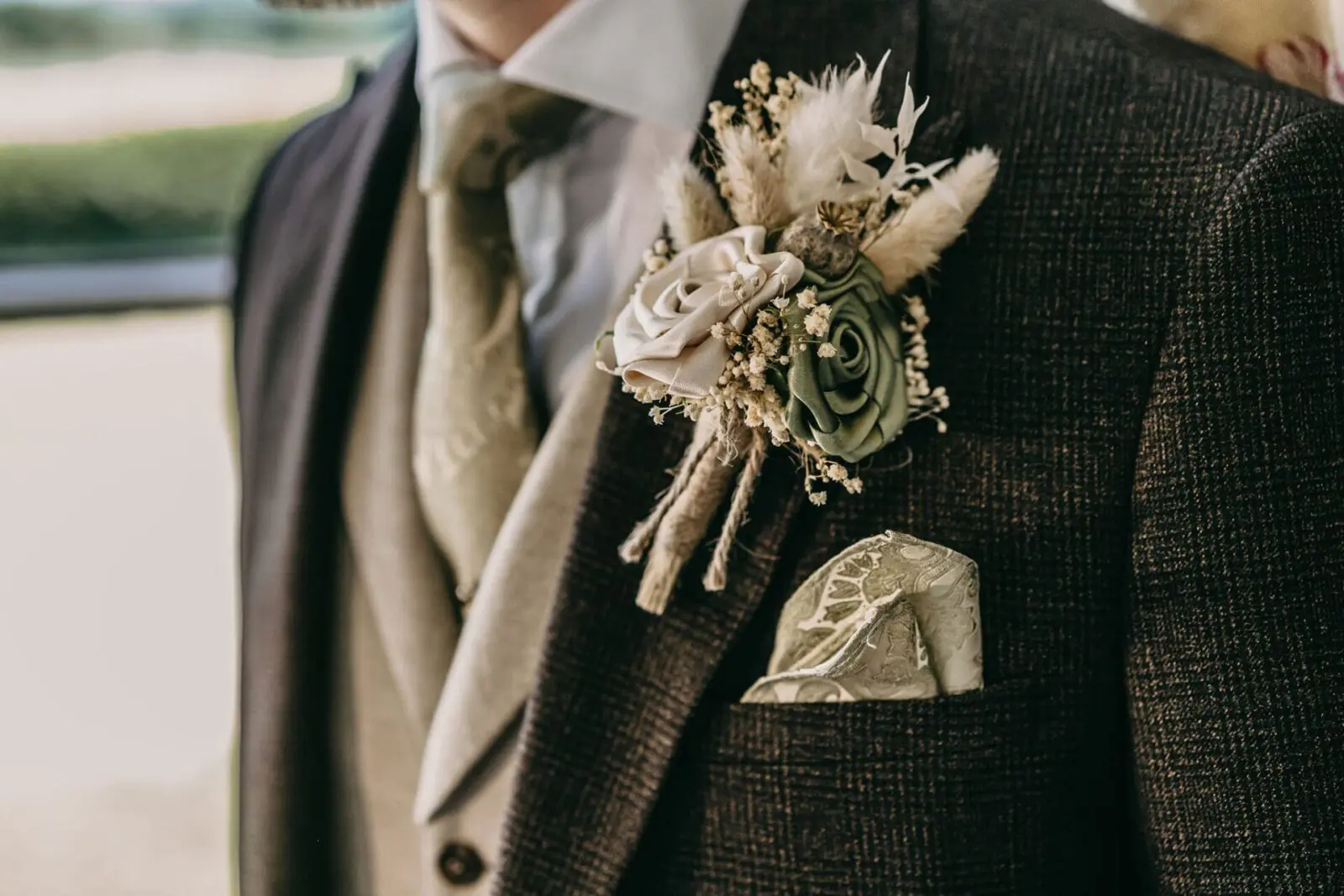 A close-up of a person wearing a brown textured suit, beige vest, patterned tie, and a faux floral arrangement boutonniere with artificial flowers and dried foliage.