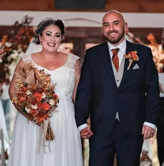 A bride in a white dress holding a bouquet and a groom in a dark suit stand side by side, smiling and holding hands indoors with fall-themed decorations.