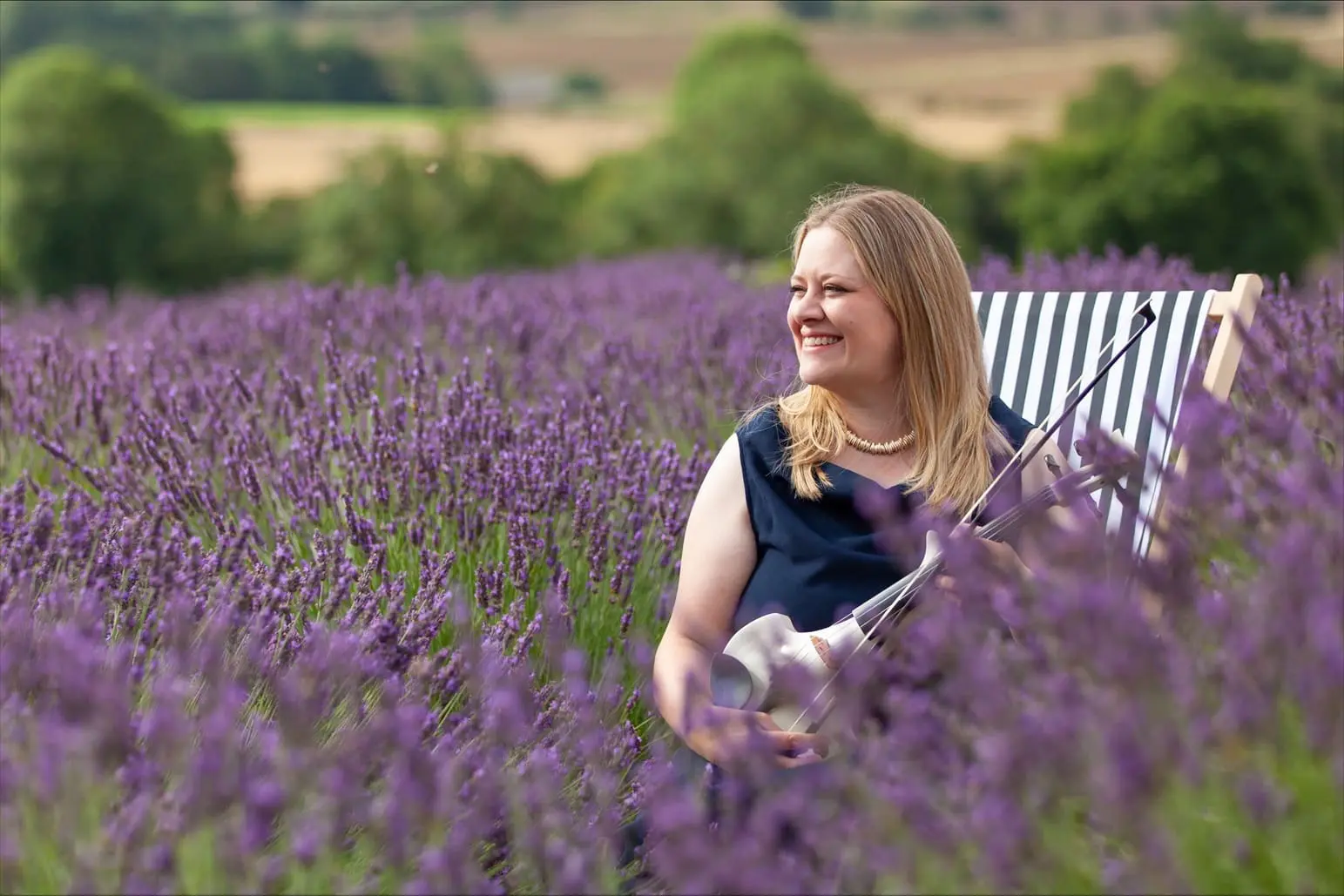 A woman in a black dress sits on a striped chair in a lavender field, holding a violin and smiling, with green trees and fields in the background.