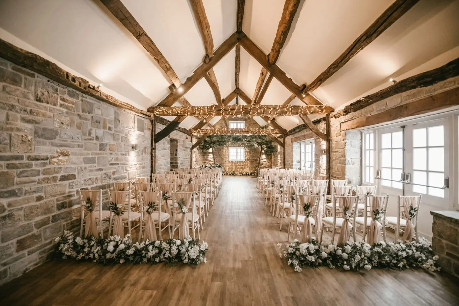 A rustic indoor wedding ceremony setup with rows of chairs, floral arrangements, exposed wooden beams, and stone walls under soft lighting.