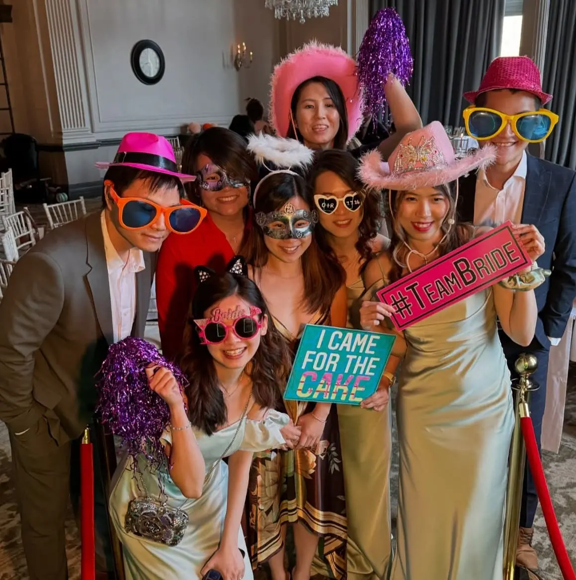 A group of people pose indoors wearing playful hats, glasses, and accessories, holding signs that read "#TeamBride" and "I Came for the Cake.