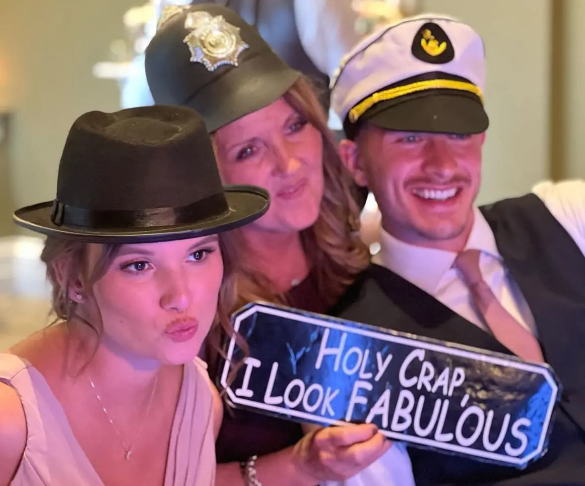Three people pose for a photo wearing different hats, with one holding a sign that reads, "Holy Crap I Look Fabulous.