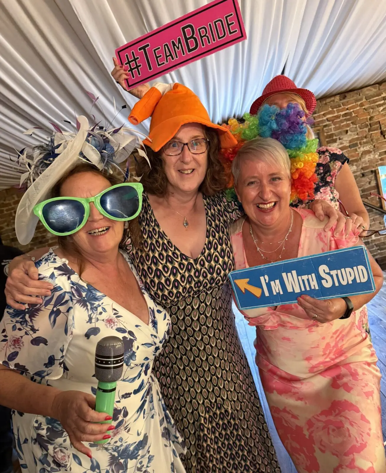 Three women at a party wear playful hats and oversized glasses, holding signs that read "#TeamBride" and "I'm With Stupid." One woman holds a microphone.