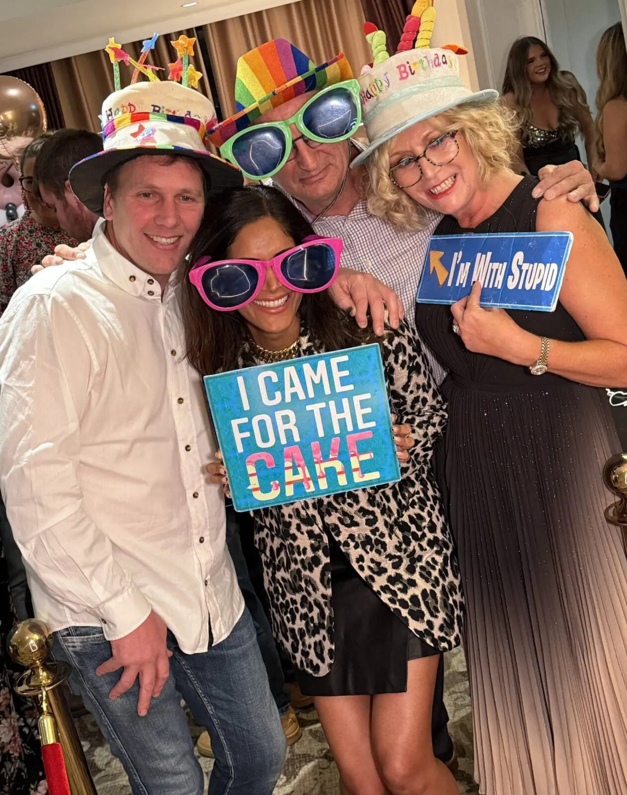 Four adults at a party wearing novelty hats and oversized glasses, holding humorous signs reading "I came for the cake" and "I'm with stupid.
