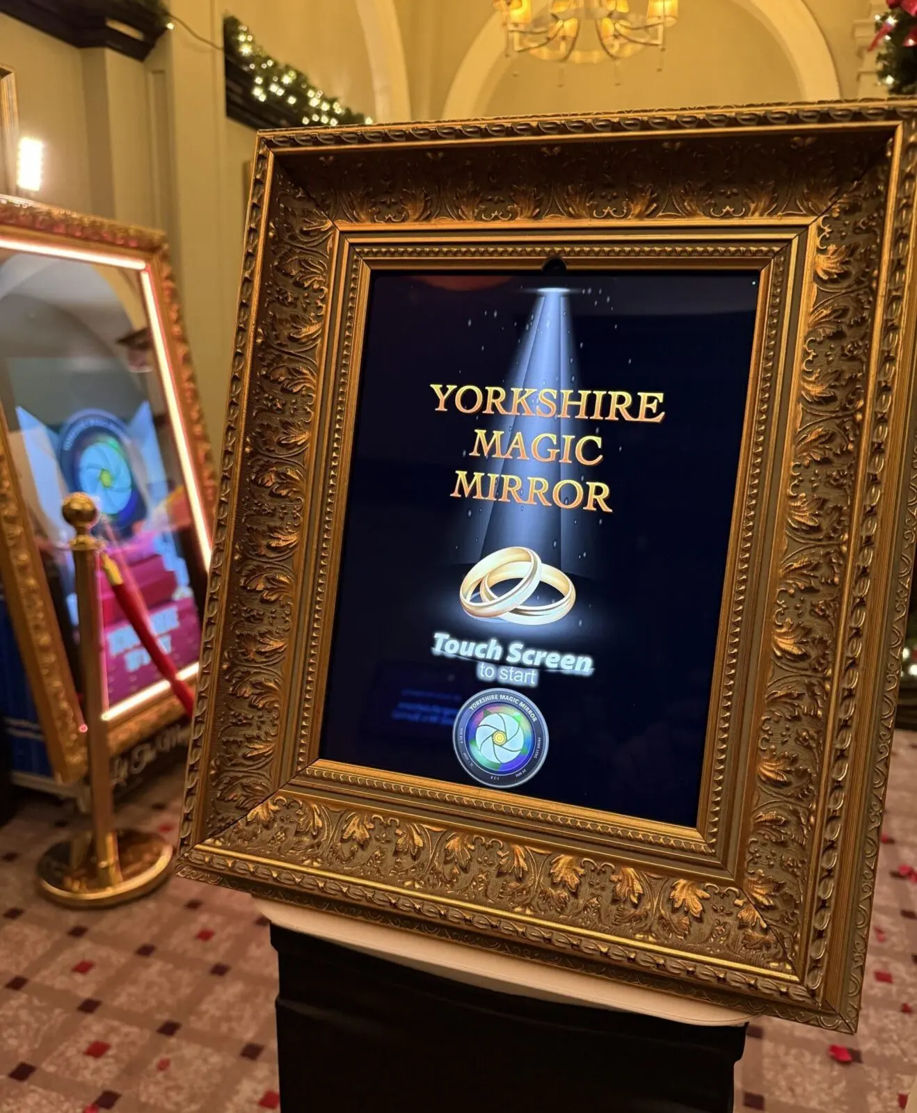 A gold ornate frame displays a magic mirror reading "Yorkshire Magic Mirror" with wedding rings and a "Touch Screen to Start" prompt in a decorated indoor setting.