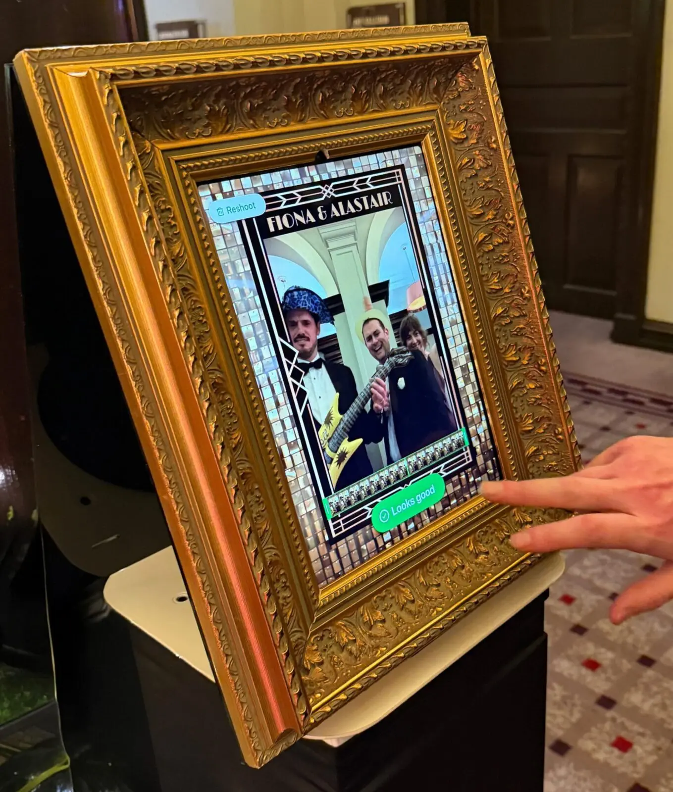 A hand touches a touchscreen magic selfie mirror display in an ornate gold frame showing a photo booth image of three people at an event.