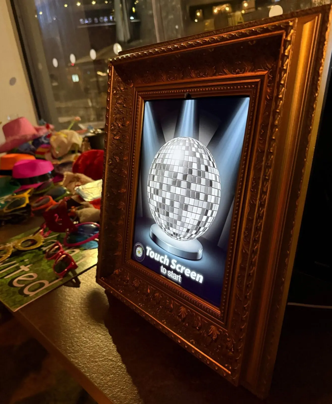 A framed digital screen displays a disco ball with the prompt "Touch Screen to start," set on a table with colorful accessories nearby.