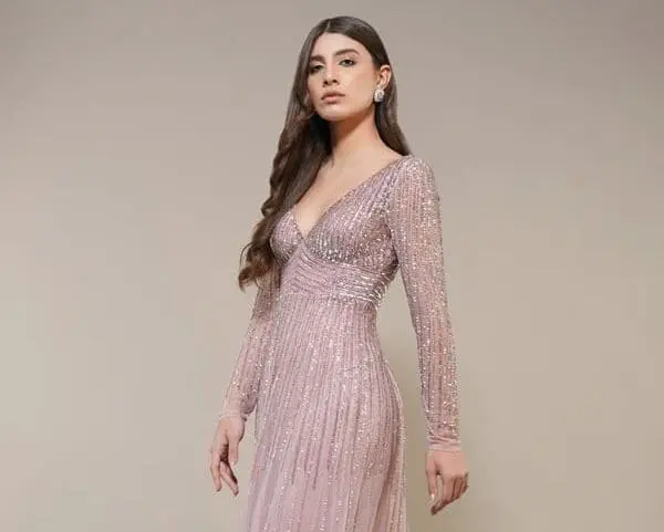 A woman wearing a long, light pink sequin gown with long sleeves and silver high-heeled sandals stands against a plain beige background.