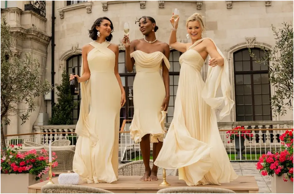 Three women in light yellow dresses stand on a platform holding champagne glasses, posing and smiling outdoors in front of a grand building with flowers and garden furniture.