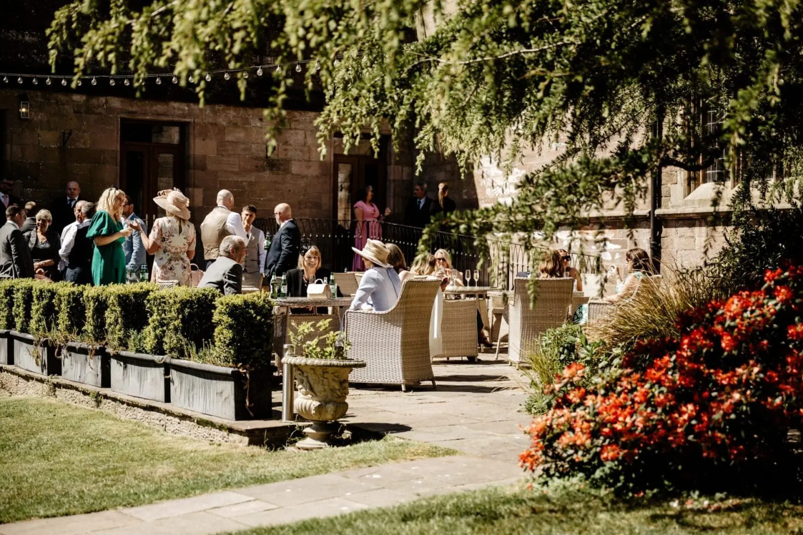 People are seated and standing in an outdoor garden area, socializing near a stone building on a sunny day. There are hedges, wicker chairs, and vibrant red flowers in the foreground.