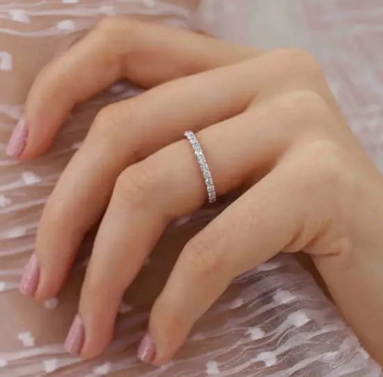 A hand with light pink polished nails wears a thin diamond eternity ring on the ring finger, resting on sheer fabric with a dotted pattern.