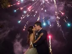 A bride and groom kiss outdoors at night whilst colourful fireworks light up the sky behind them.