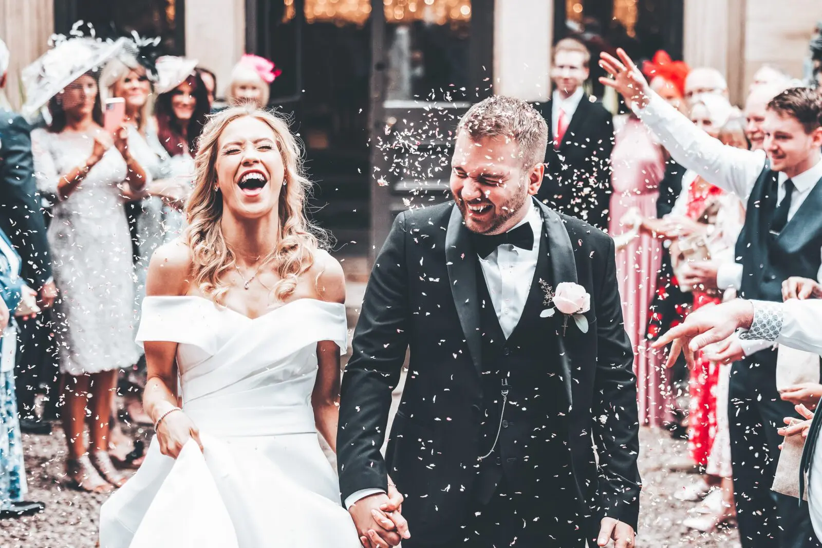 A bride and groom, both smiling, walk outdoors holding hands whilst guests throw confetti around them at a wedding celebration.