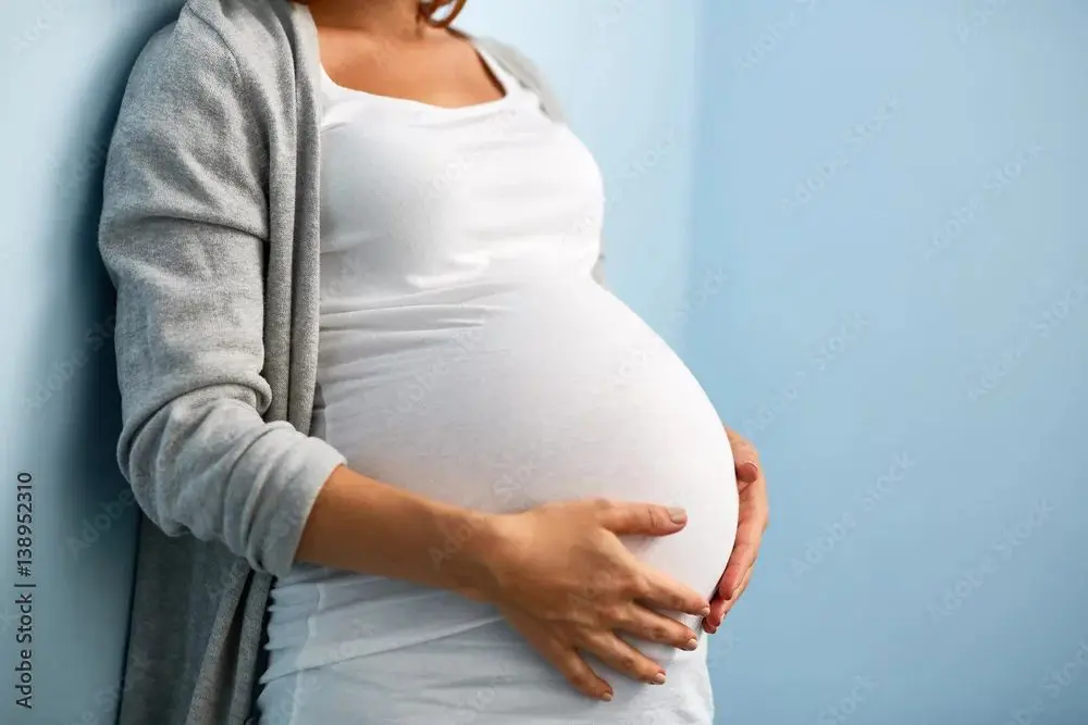 Pregnant woman in a white top and grey cardigan leaning against a blue wall, holding her belly with both hands.