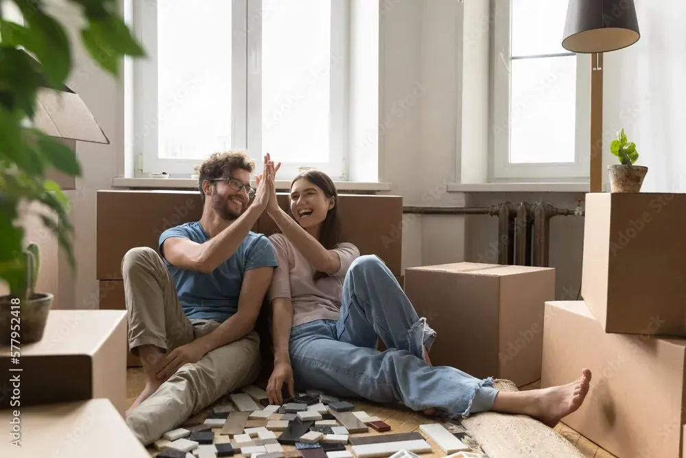 A smiling couple sits on the floor among moving boxes, giving each other a high five, with tiles spread out in front of them in a bright room.