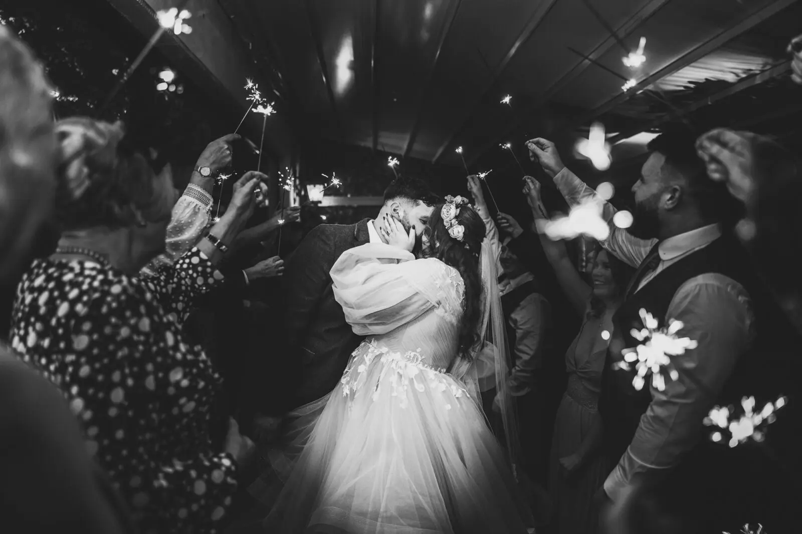 A bride and groom kiss surrounded by guests holding sparklers during a wedding celebration.