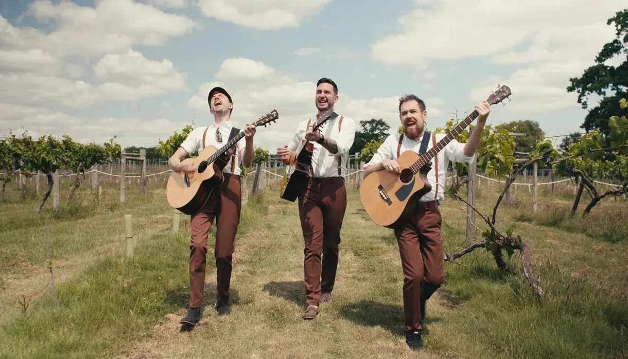 Three men wearing white shirts, brown trousers, and braces walk through a vineyard playing acoustic guitars and singing under a partly cloudy sky.