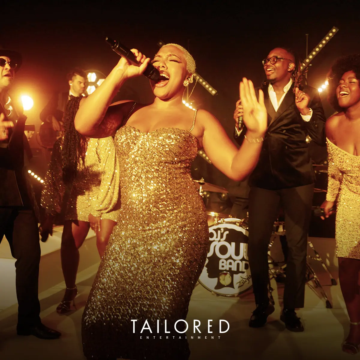 A band performs on stage under warm lighting, with a lead singer in a gold dress holding a microphone and other members singing and dancing.