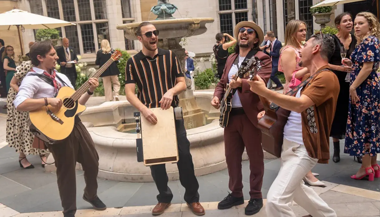 Four musicians perform energetically with guitars and a wooden percussion instrument in a courtyard, surrounded by people socialising near a fountain.