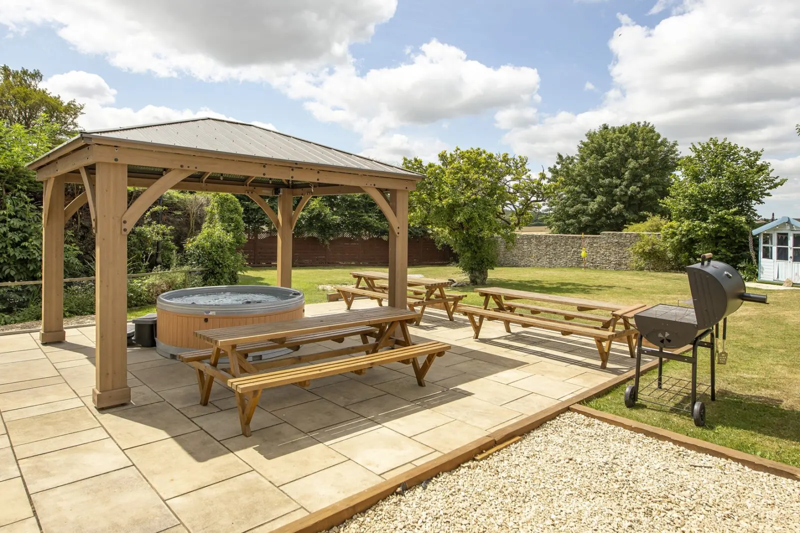 Wooden gazebo with a hot tub, picnic tables, and a barbecue on a paved patio in a sunny garden with trees and a shed in the background.