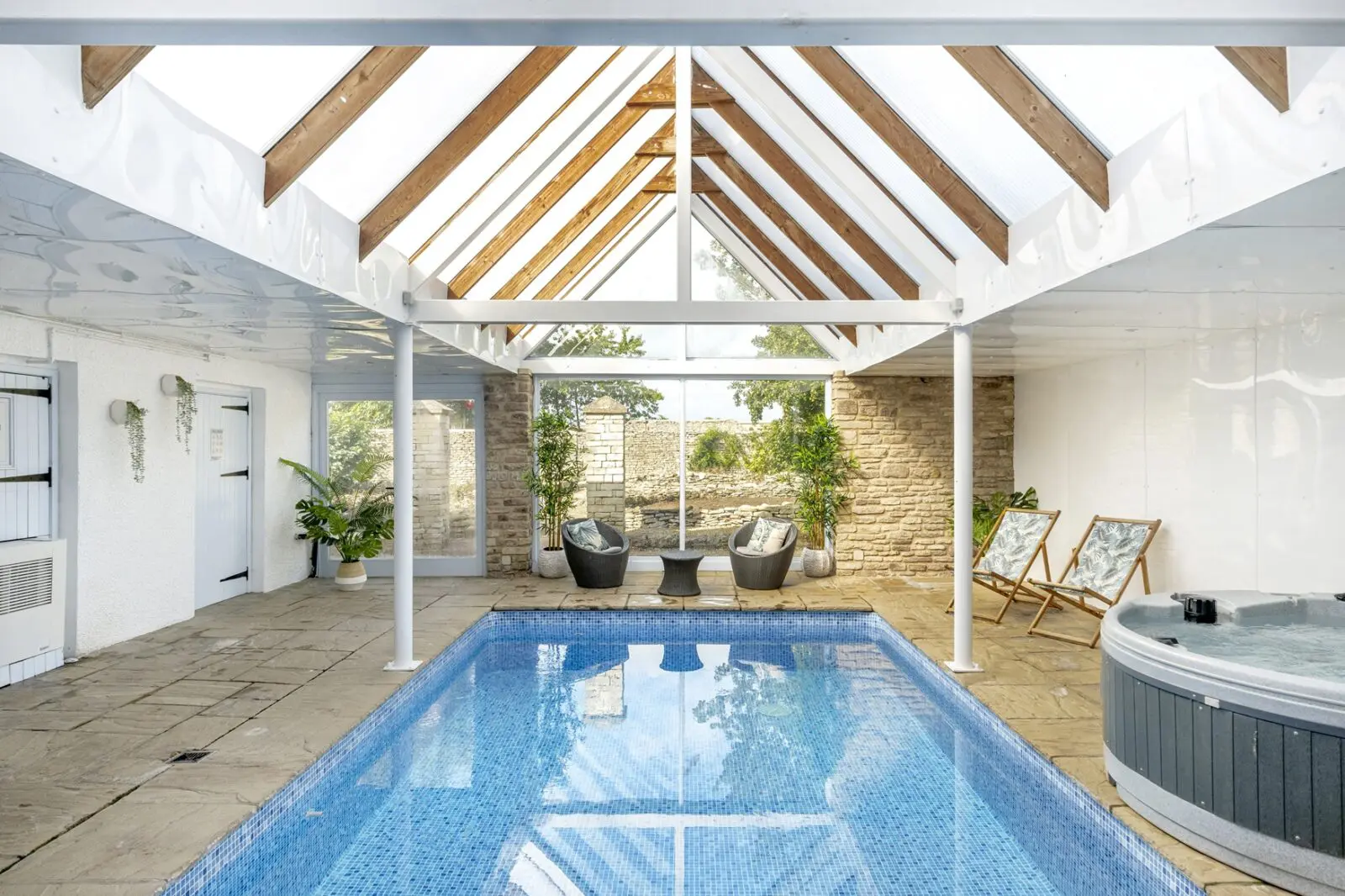 Indoor pool room with a blue-tiled pool, hot tub, lounge chairs, potted plants, stone wall, and glass ceiling letting in natural light.