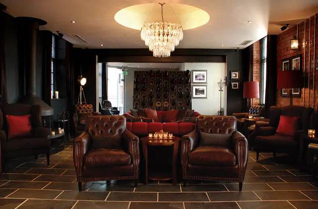 A lounge with leather armchairs, a settee, a round chandelier, dim lighting, dark tiled floors, and framed art on exposed brick walls.